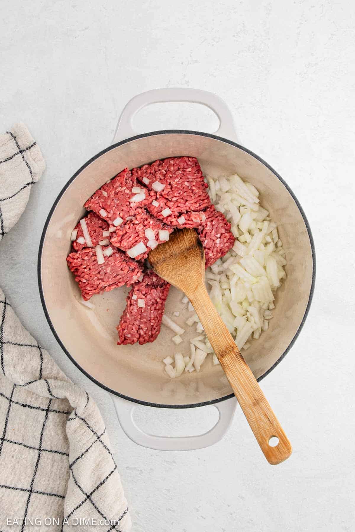 A white pot on a light surface contains raw ground beef and chopped onions, essential ingredients for a hearty hamburger soup. A wooden spoon rests inside the pot, ready to stir, while a checked cloth is partly visible on the left side.