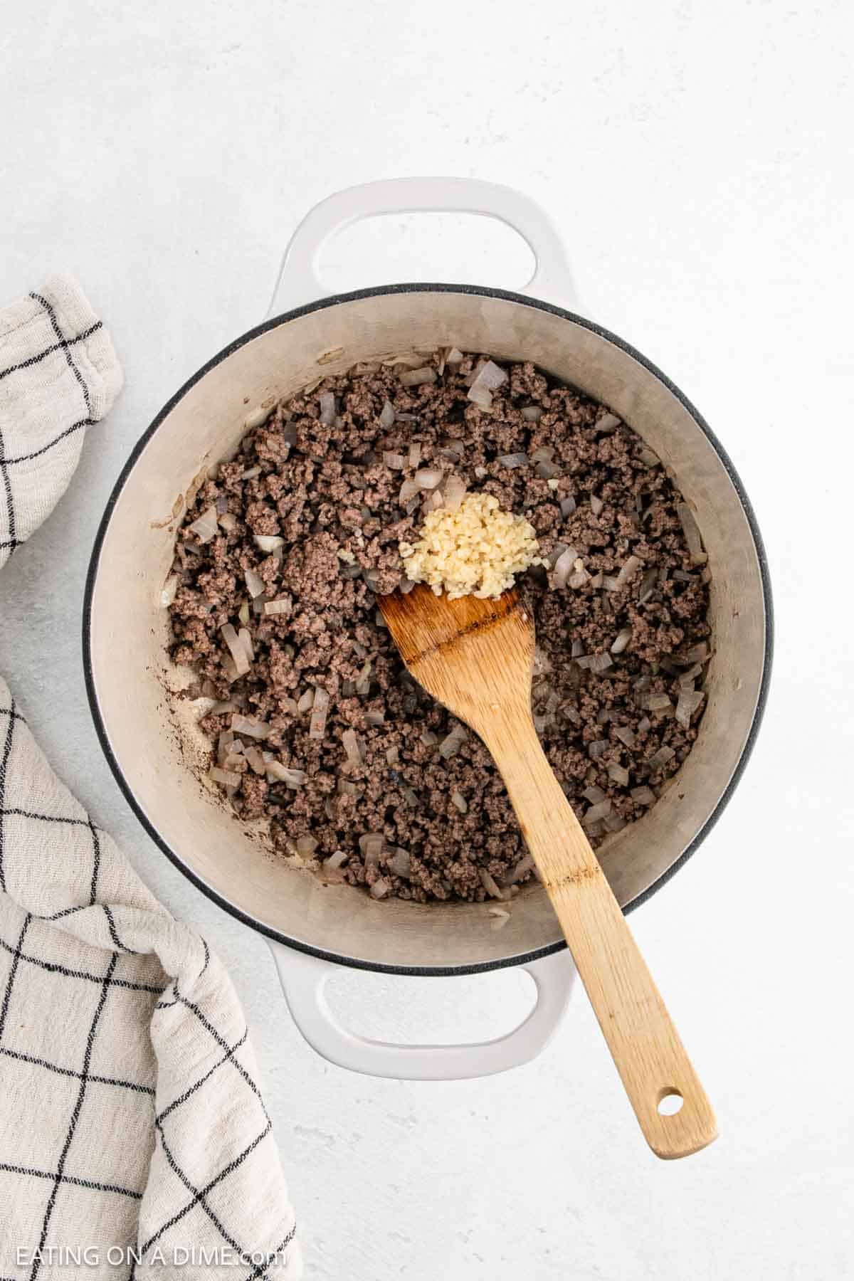 A white pot filled with cooked ground beef and onions sits ready for transformation into a comforting Hamburger Soup. Minced garlic is added on top, and a wooden spoon rests inside. A black and white checkered kitchen towel lies beside it on a light-colored surface, setting the cozy scene.