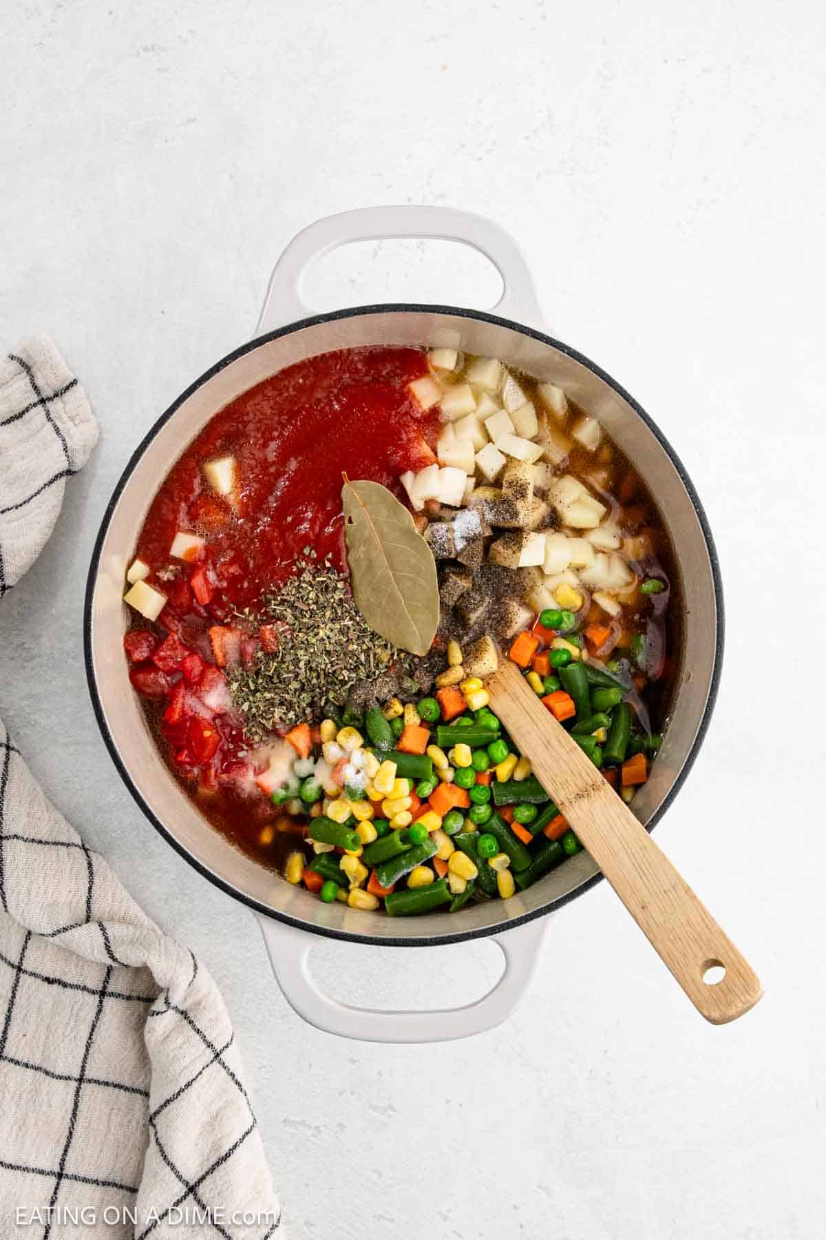 A pot of hearty vegetable soup ingredients on a light surface. The recipe includes tomato sauce, diced tomatoes, potatoes, mixed vegetables, seasonings, and a bay leaf. A wooden spoon rests inside the pot. A plaid kitchen towel lies to the side. Perfect for pairing with hamburger crumbles for extra flavor!.