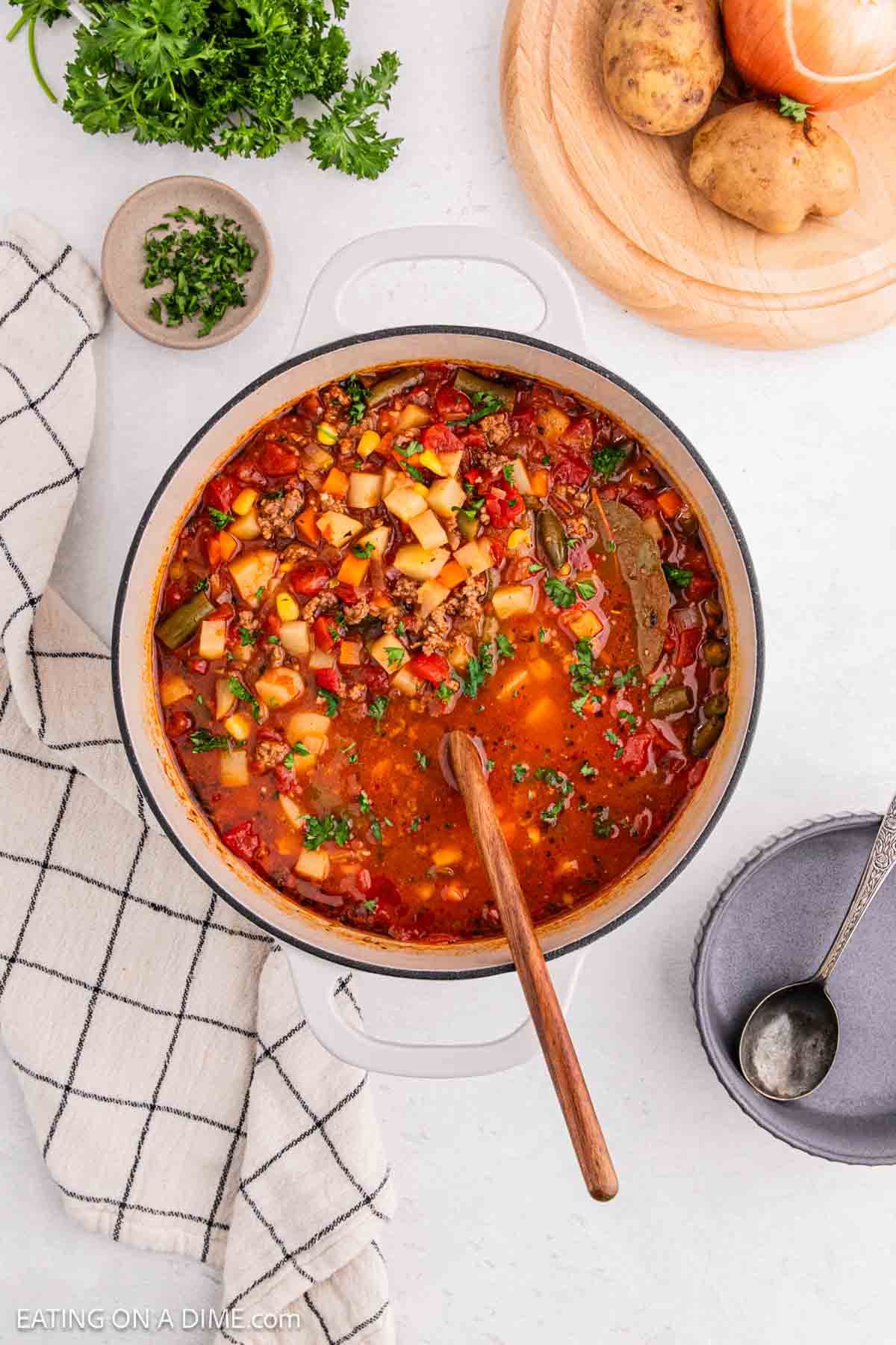 A pot of hearty vegetable soup, reminiscent of a classic hamburger, with chopped herbs on top, surrounded by a kitchen towel, an empty bowl with a spoon, and a wooden board holding potatoes and an onion. This delicious recipe brims with diced vegetables in a rich broth.