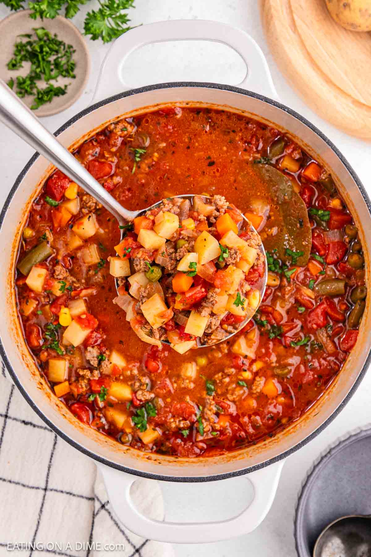 A pot of hearty hamburger soup filled with beef, diced potatoes, tomatoes, and various vegetables is shown. A ladle lifts a serving, displaying chunks of vegetables and meat. Fresh herbs are visible on the side, enhancing the presentation.