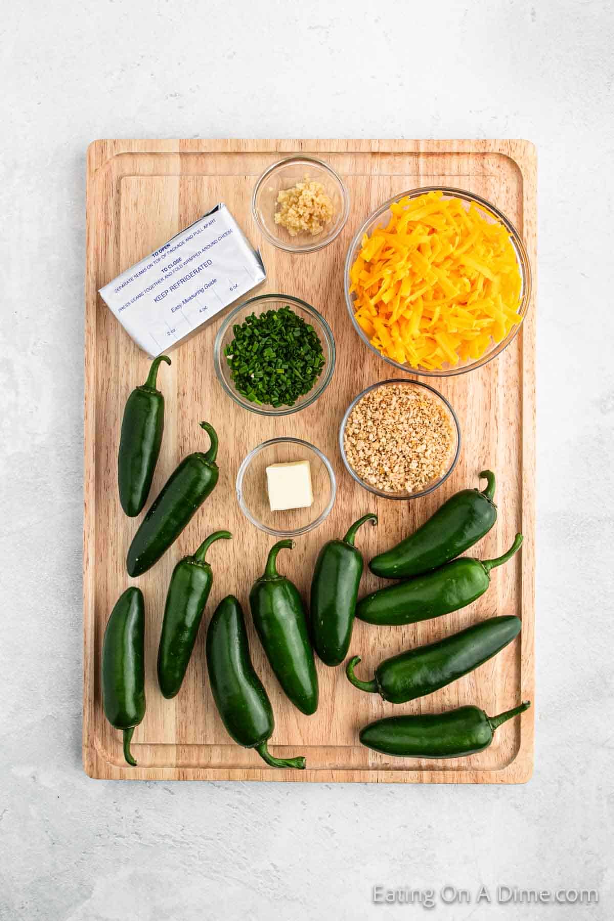 A wooden cutting board showcases jalapeño poppers essentials: fresh jalapeños, a block of cream cheese, grated cheddar cheese, minced garlic, chopped herbs, sesame seeds, and a small piece of butter—ideal for crafting spicy snacks that'll heat up any gathering.