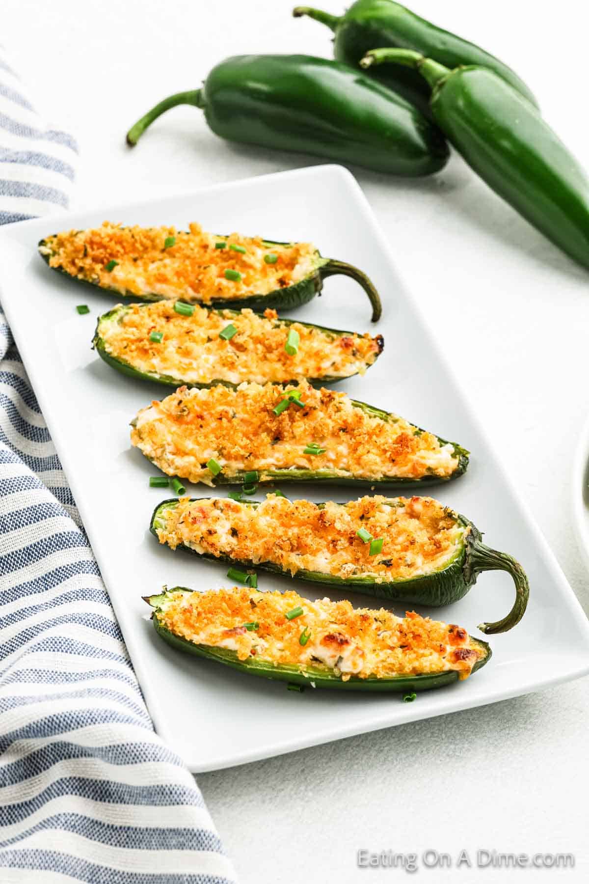 A plate of six jalapeño poppers filled with melted cheese and crispy breadcrumbs, garnished with chopped herbs. Two fresh jalapeños and a striped towel are in the background, perfectly highlighting these irresistible snacks.