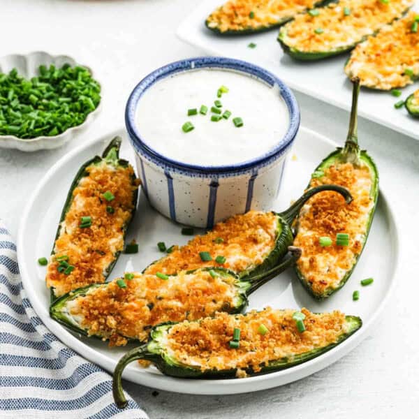 A plate of jalapeño poppers topped with breadcrumbs and chives surrounds a bowl of creamy dipping sauce. Fresh chives are in a small dish nearby, while a striped napkin peeks from the edge, inviting you to savor every spicy bite.