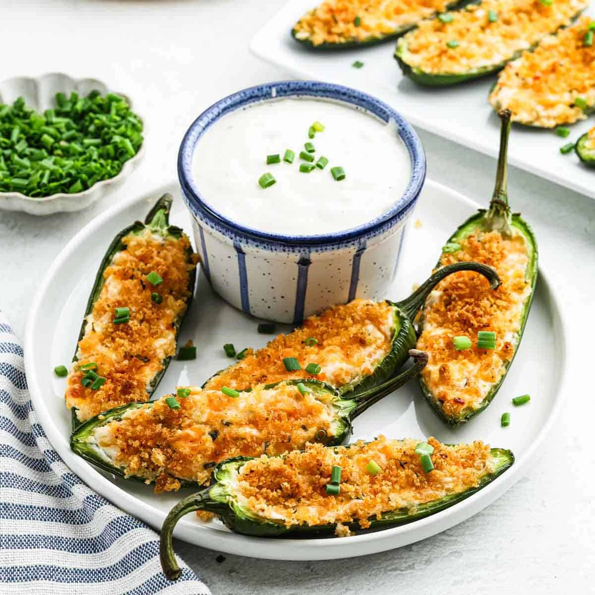 A plate of jalapeño poppers topped with breadcrumbs and chives surrounds a bowl of creamy dipping sauce. Fresh chives are in a small dish nearby, while a striped napkin peeks from the edge, inviting you to savor every spicy bite.