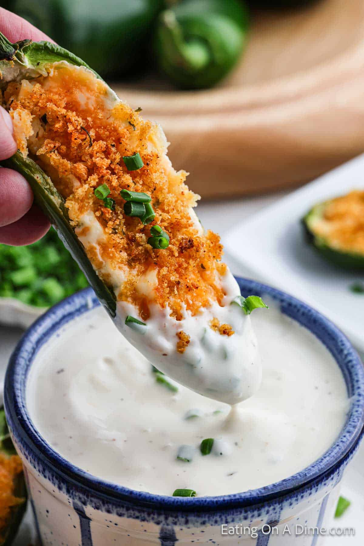 A jalapeño popper, the star of spicy snacks and appetizers, is dipped into creamy white sauce. The popper oozes with a cheesy filling and is crowned with crispy breadcrumbs and chopped green onions. More tempting jalapeños and a bowl of sauce wait enticingly in the background.