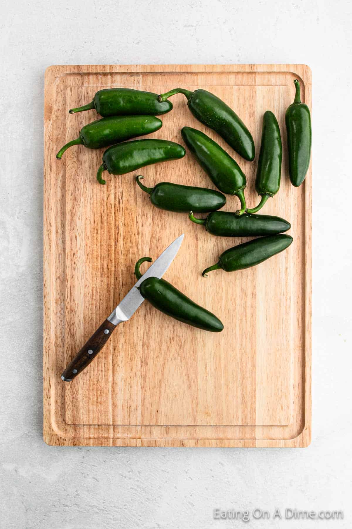 A wooden cutting board holds about a dozen green jalapeños and a small knife, perfect for preparing spicy Jalapeño Poppers as an appetizer. The board rests on a light gray surface, with the jalapeños mostly arranged in the upper right corner.