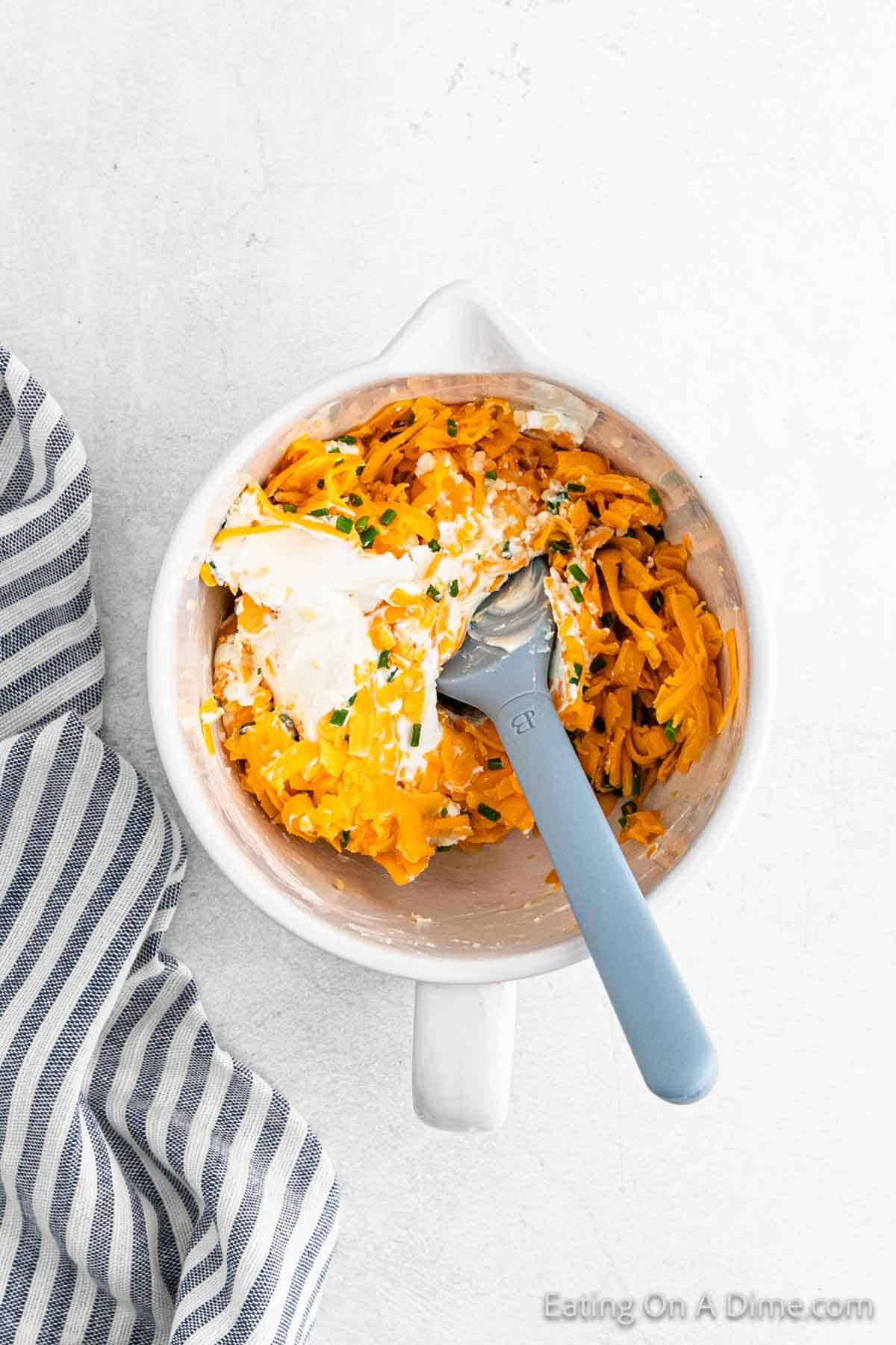 A white bowl brimming with a jalapeño poppers-inspired mix of shredded cheddar cheese, cream cheese, and chopped green herbs is stirred with a blue spoon. The inviting bowl nestles beside a striped cloth on a light surface.