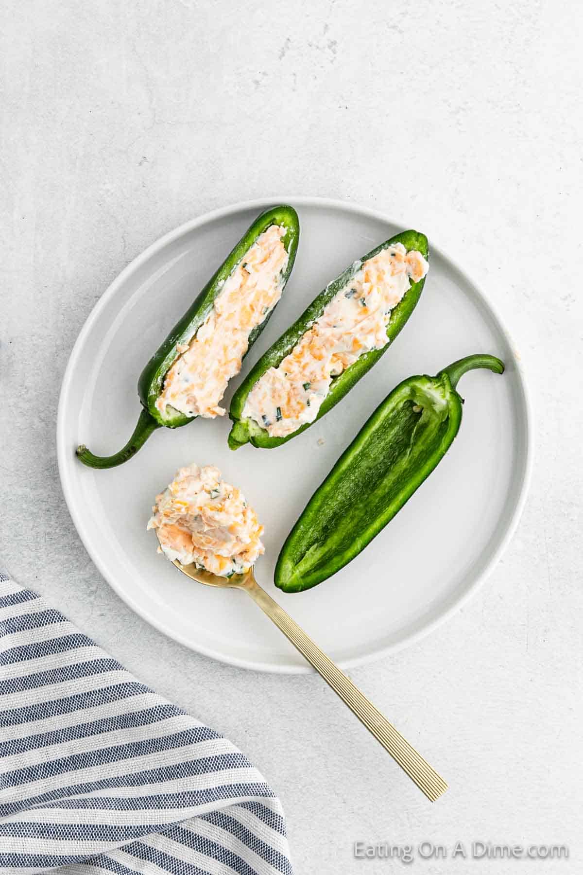 A white plate with Jalapeño Poppers halved and filled with a creamy cheese mixture. One half is on a spoon, resting on the plate. A gray and white striped cloth is nearby on the table, completing this perfect spicy snack setting.