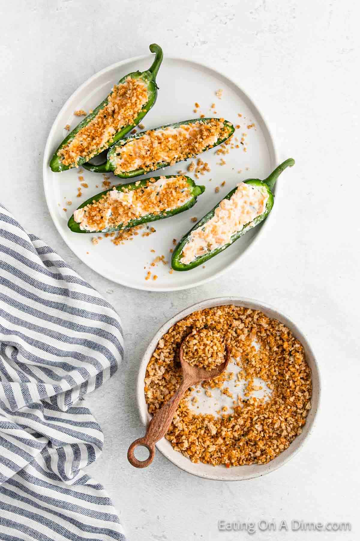 A plate of jalapeño poppers filled with a creamy mixture and topped with breadcrumbs sits next to a bowl of crunchy breadcrumbs with a wooden spoon. A striped cloth adds charm to the light surface, completing the spicy appetizer's enticing presentation.