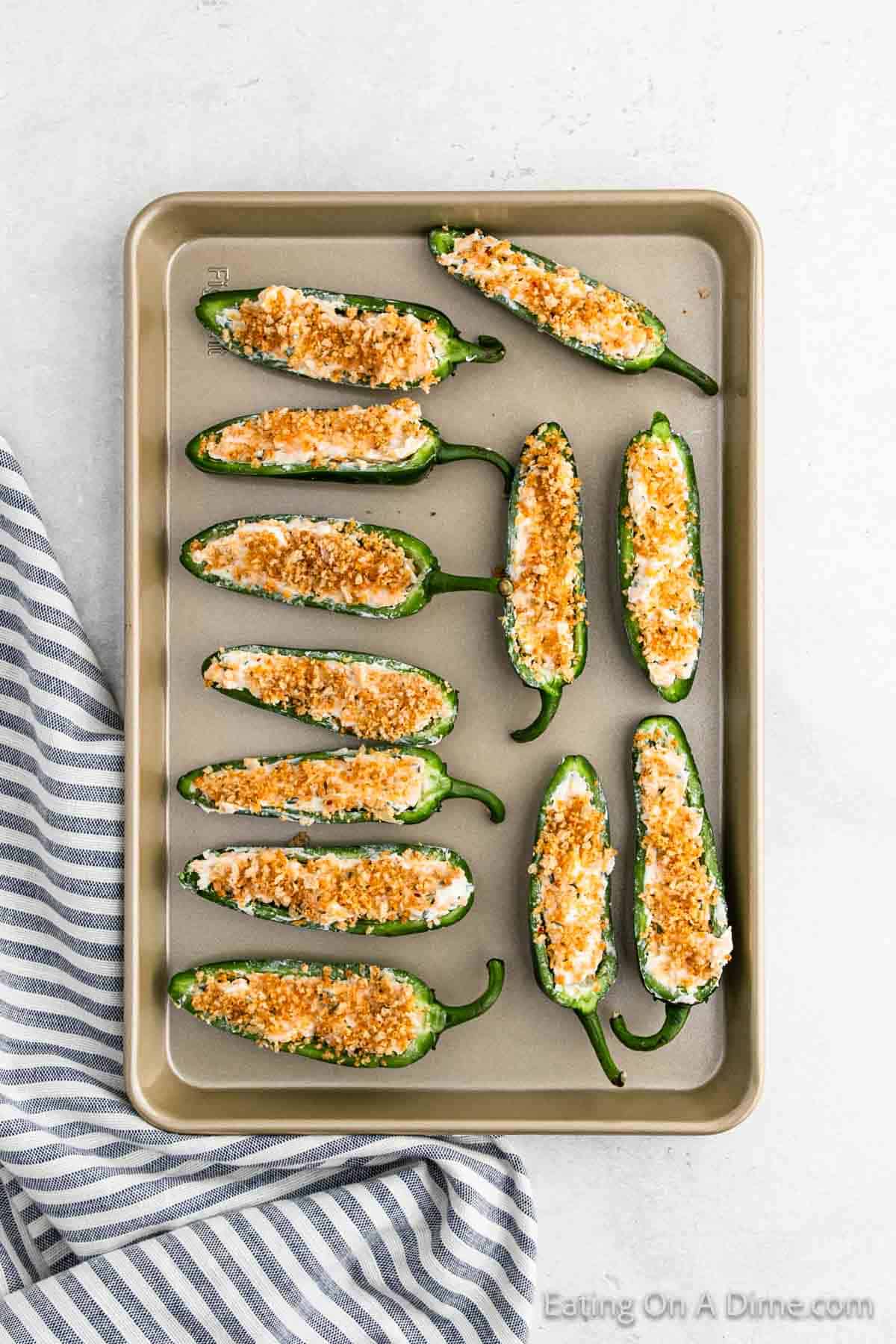 A baking tray with jalapeño poppers, featuring halved jalapeños filled with cream cheese and topped with breadcrumbs. This savory delight rests on a light surface next to a striped cloth.