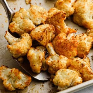 A close-up of roasted cauliflower florets on a baking sheet, perfectly seasoned with spices. One piece is being scooped up with a metal spatula, showcasing its golden-brown, crispy appearance.