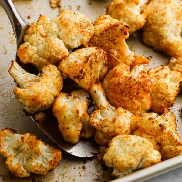 A close-up of roasted cauliflower florets on a baking sheet, perfectly seasoned with spices. One piece is being scooped up with a metal spatula, showcasing its golden-brown, crispy appearance.