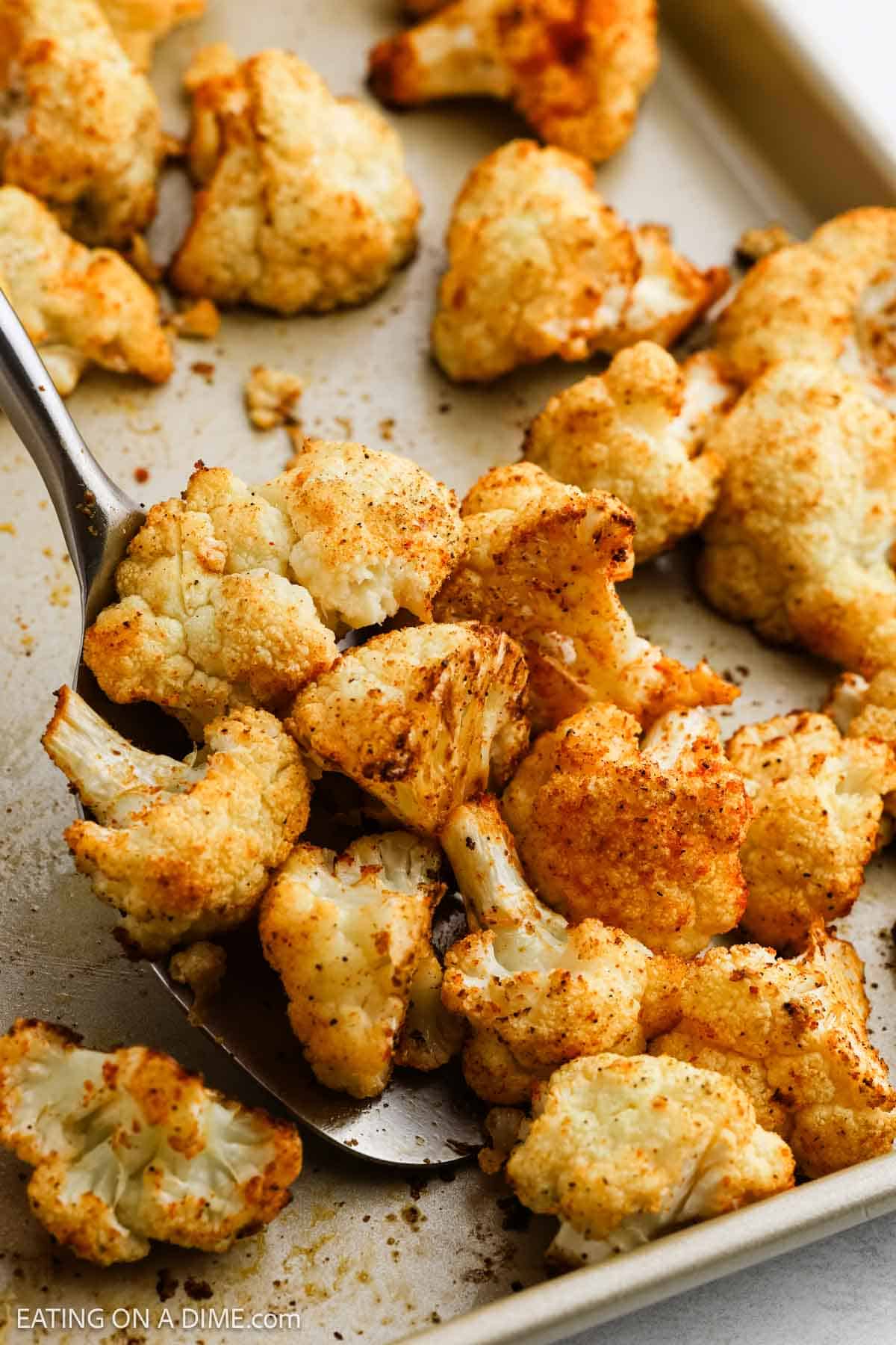 A baking sheet with roasted cauliflower florets seasoned to perfection. A spatula lifts some of them, revealing a golden-brown, crispy texture. The roasted cauliflower is sprinkled with spices, giving it a savory appearance.