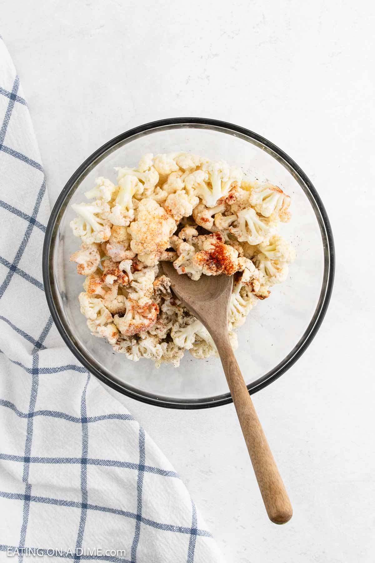 A glass bowl brimming with roasted cauliflower, perfectly seasoned with spices, sits invitingly. A wooden spoon rests inside, ready for serving. Beside the bowl lies a blue-and-white checkered cloth on a light surface, adding a touch of homely charm.