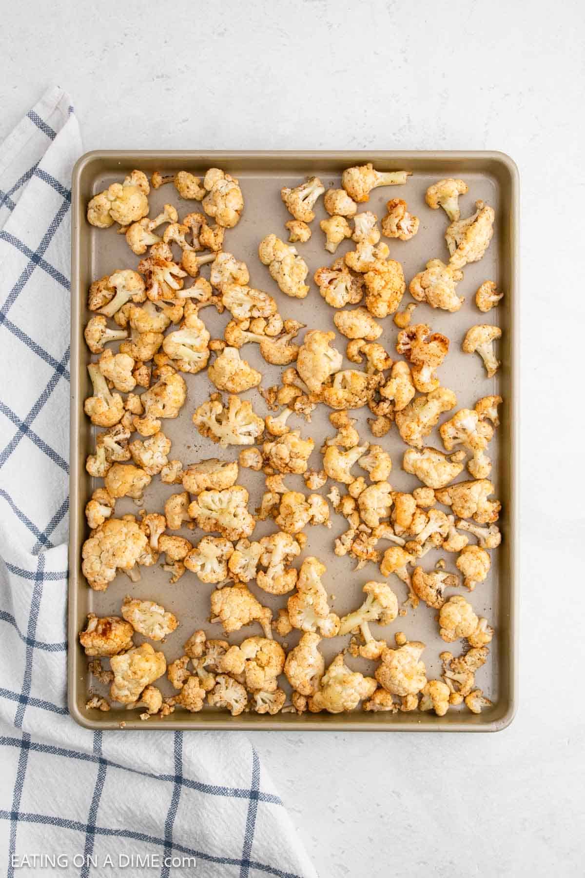 A baking sheet brimming with roasted cauliflower florets, golden-brown and perfectly seasoned. The sheet rests on a white surface with a blue-checkered kitchen towel partially visible to the left.