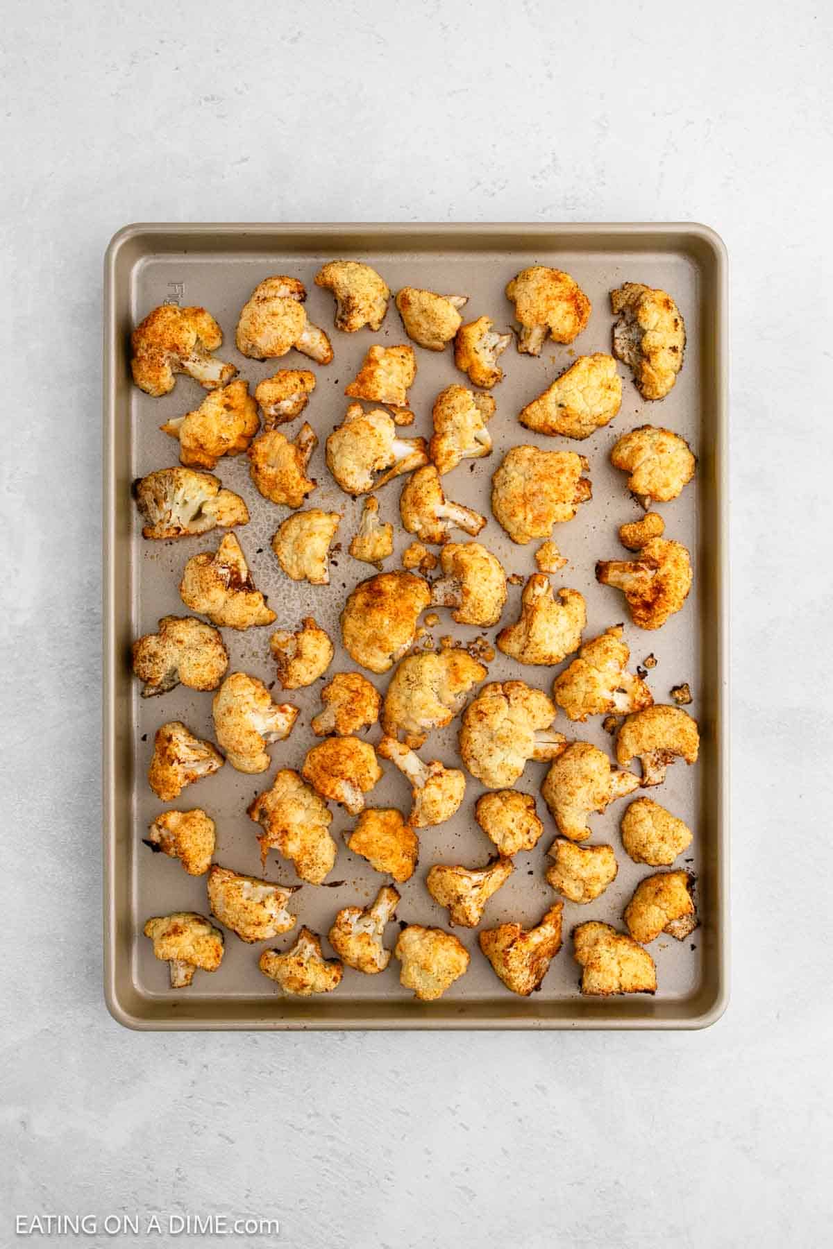 Golden-brown roasted cauliflower florets are artfully arranged on a baking sheet, lightly seasoned to perfection. This culinary display rests elegantly on a textured light gray surface, highlighting the simplicity and beauty of roasted cauliflower.