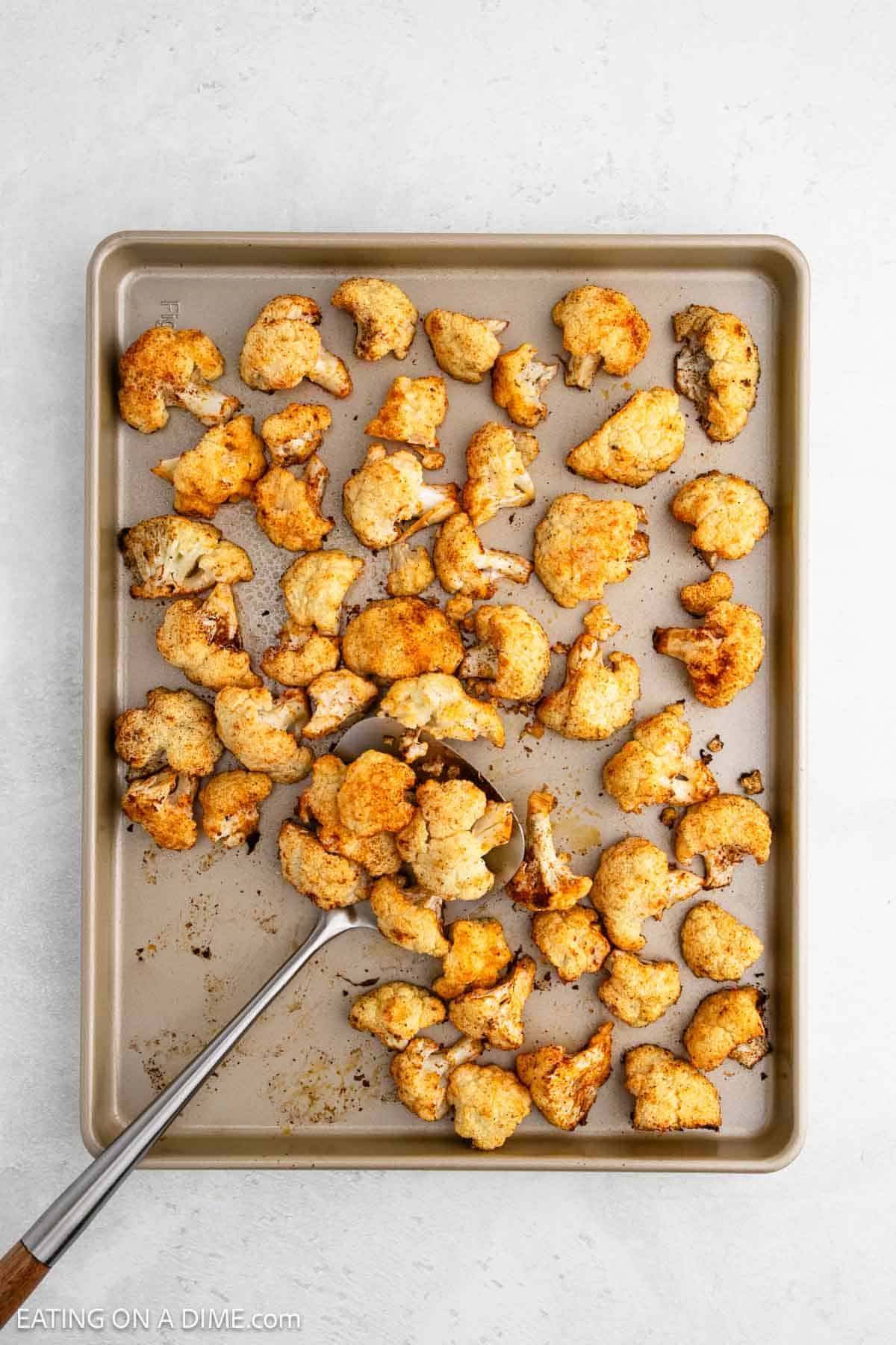 Golden brown roasted cauliflower pieces, perfectly seasoned with spices, lie enticingly on a baking sheet next to a serving spoon. The light gray surface in the background complements the enticing dish.