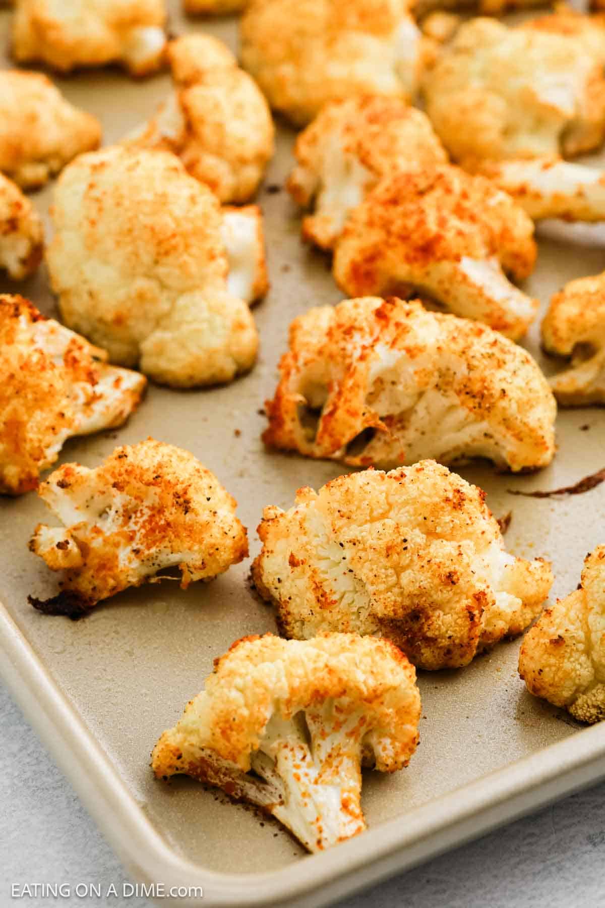 Close-up of roasted cauliflower florets on a baking tray. These roasted cauliflower pieces are seasoned with spices, giving them a golden, crispy appearance. The tray rests on a light-colored surface, and the florets are evenly spread.