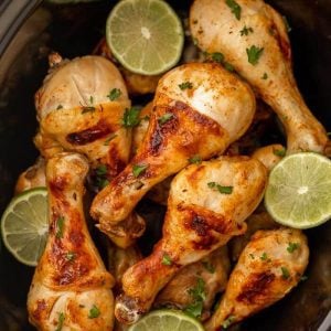 Grilled chicken drumsticks garnished with chopped herbs are piled together, surrounded by lime slices. This delicious dish from Meal Plan 42 features golden brown, juicy chicken in a dark dish, perfect for your dining table.