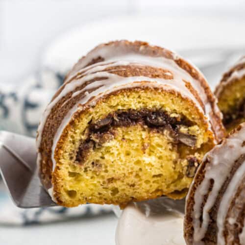 Close-up of a sliced Sock It To Me Cake reveals its moist, golden interior with a rich chocolate filling. The cake is topped with a light glaze drizzle and rests elegantly on a cake server, ready to delight taste buds.