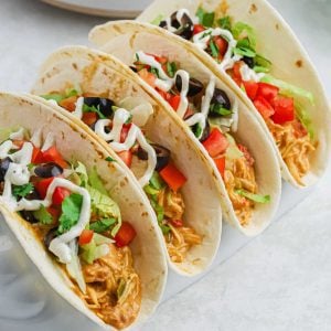 Four soft tacos are filled with shredded chicken, diced tomatoes, black olives, lettuce, and cilantro. They are topped with a drizzle of white sauce and neatly arranged on a taco holder—a perfect addition to your Meal Plan 45.