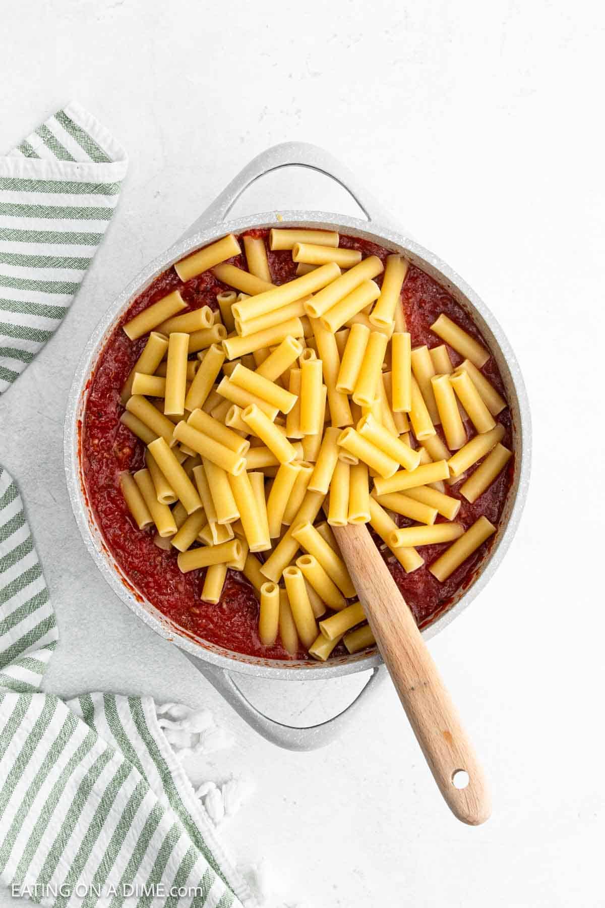 Cooked Ziti Pasta topped with pasta sauce mixture in a large skillet with a wooden spoon