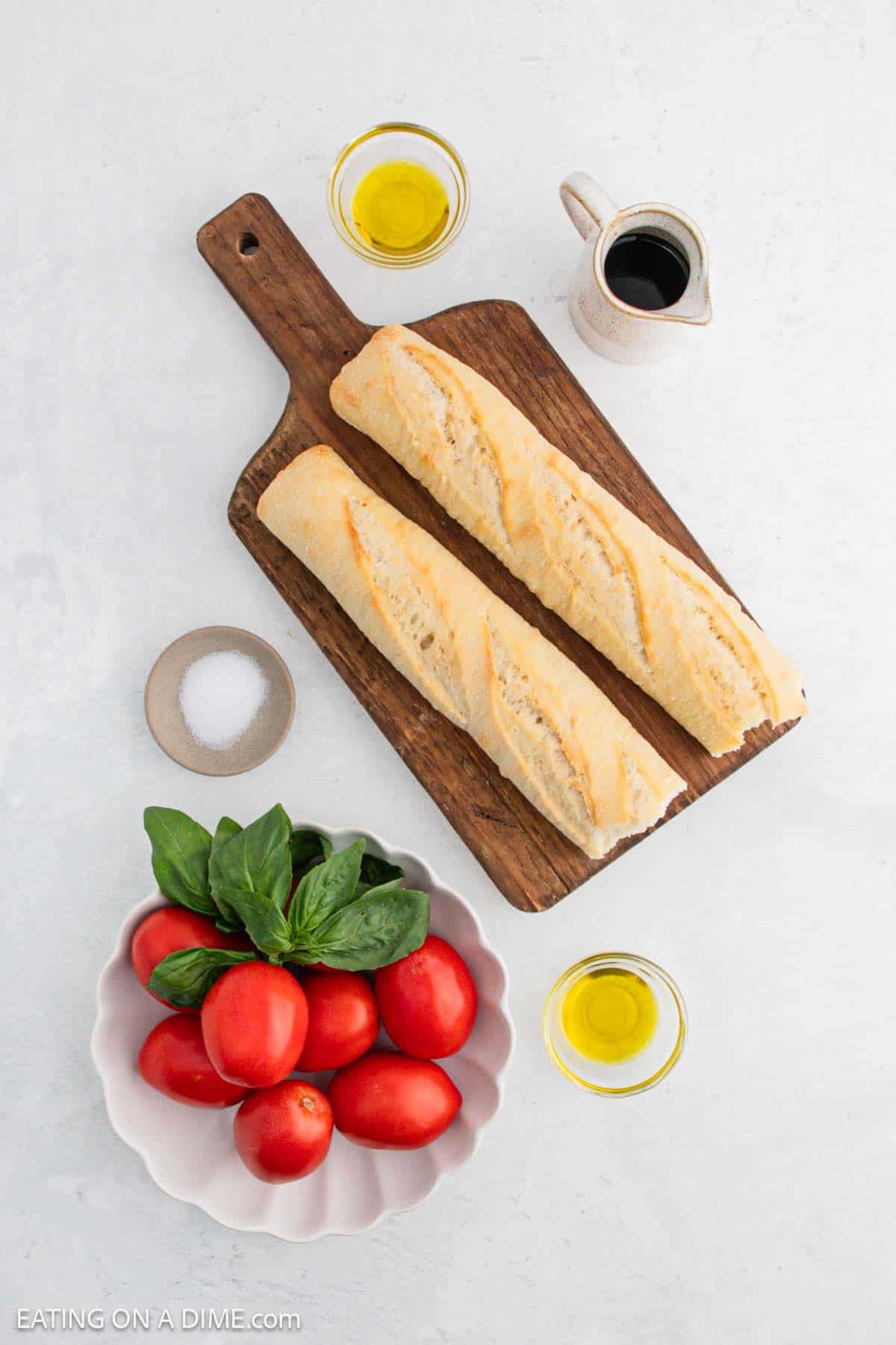 Two baguettes on a wooden cutting board, surrounded by a small pitcher, bowls of olive oil, a dish with tomatoes and basil, and a bowl of salt. The setting is on a white surface.