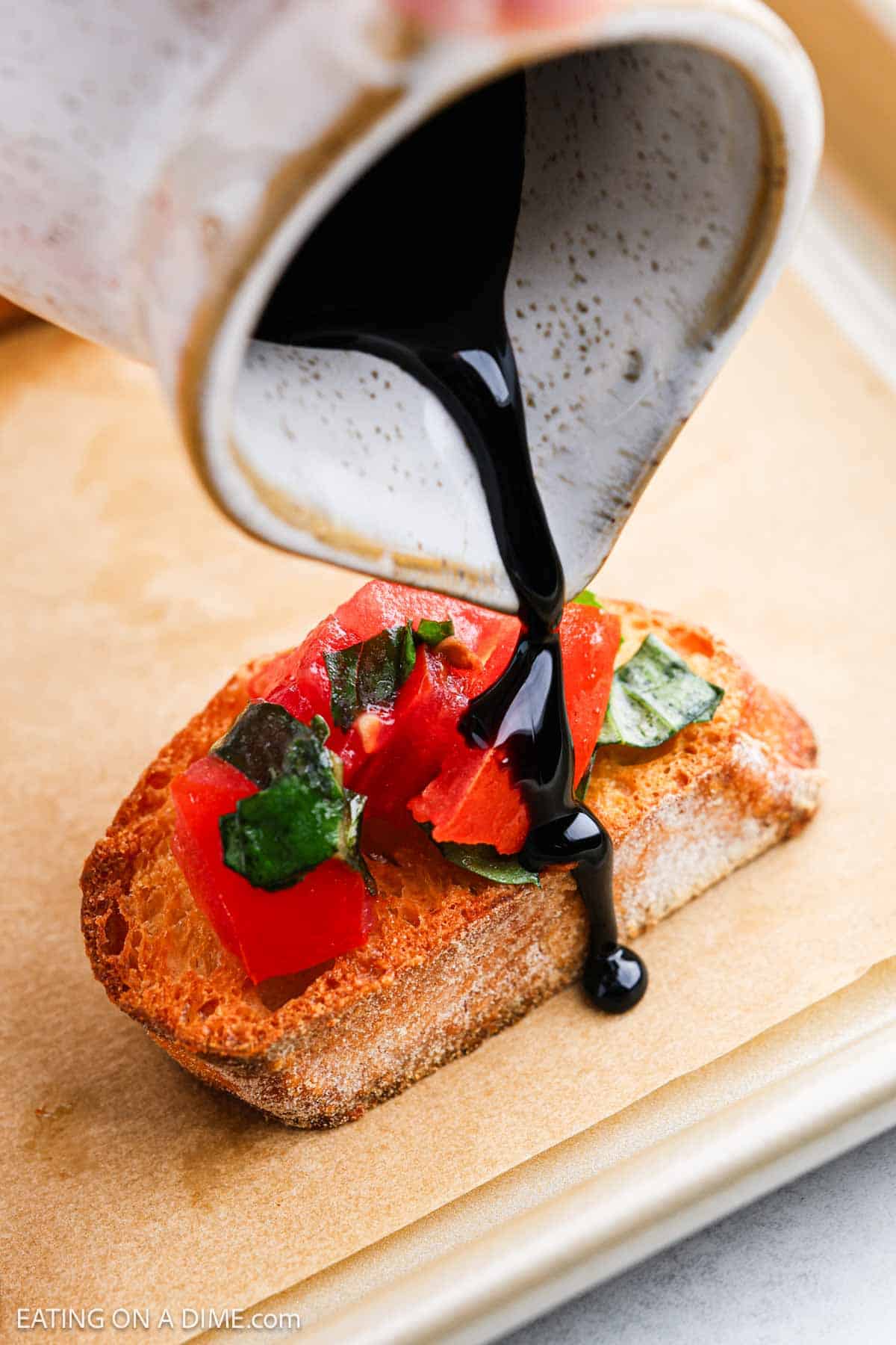 A close-up of balsamic glaze being poured from a small pitcher over a bruschetta topped with diced tomatoes and fresh basil on toasted bread. The image is set on a parchment-lined baking sheet.