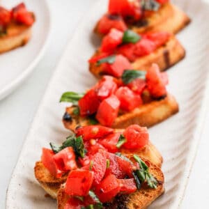 A plate of bruschetta topped with diced tomatoes and fresh basil on toasted slices of bread. Each serving is arranged in a row, showcasing the vibrant red and green colors against the golden brown toast.