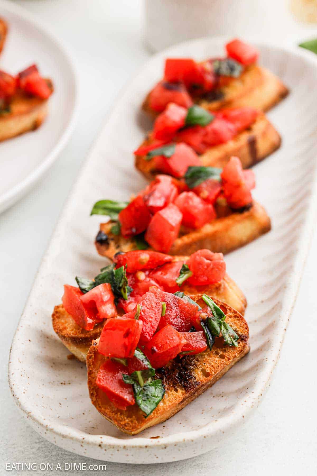 A plate of bruschetta topped with diced tomatoes, chopped basil, and olive oil on toasted bread slices. The bruschetta is served on an elongated white ceramic dish.