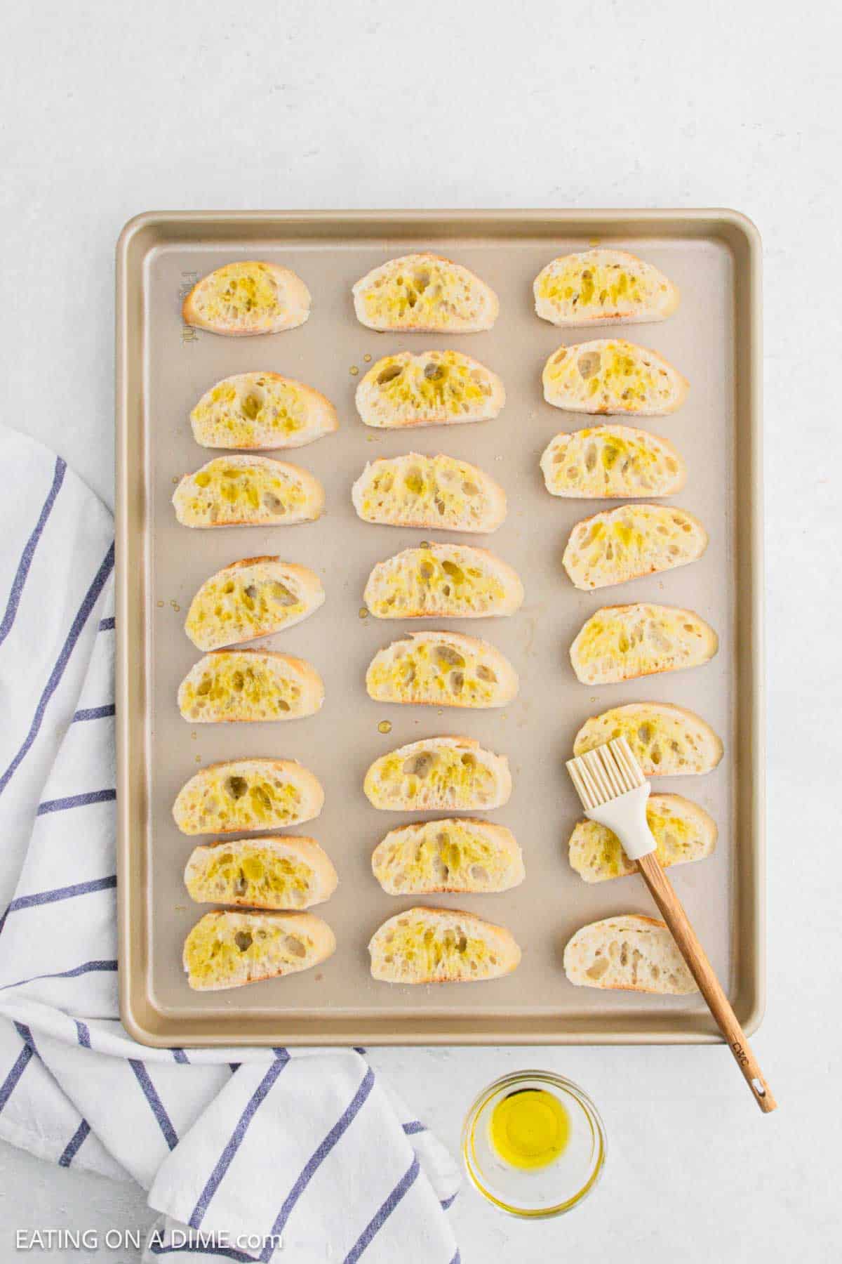 A baking sheet with sliced bread pieces topped with olive oil, arranged neatly. A brush rests on the sheet, and a small bowl of olive oil is nearby. A striped towel is partially visible on the left side.