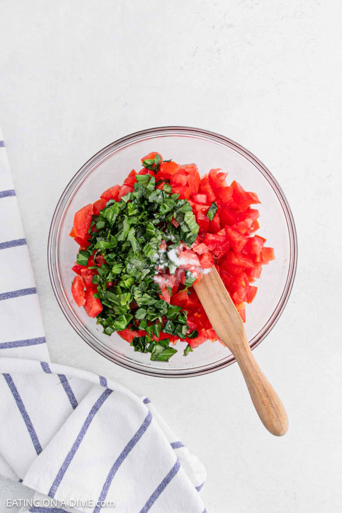 A glass bowl filled with chopped tomatoes, green herbs, and a sprinkle of salt, with a wooden spoon inside. The bowl is on a white surface next to a white and blue striped cloth.