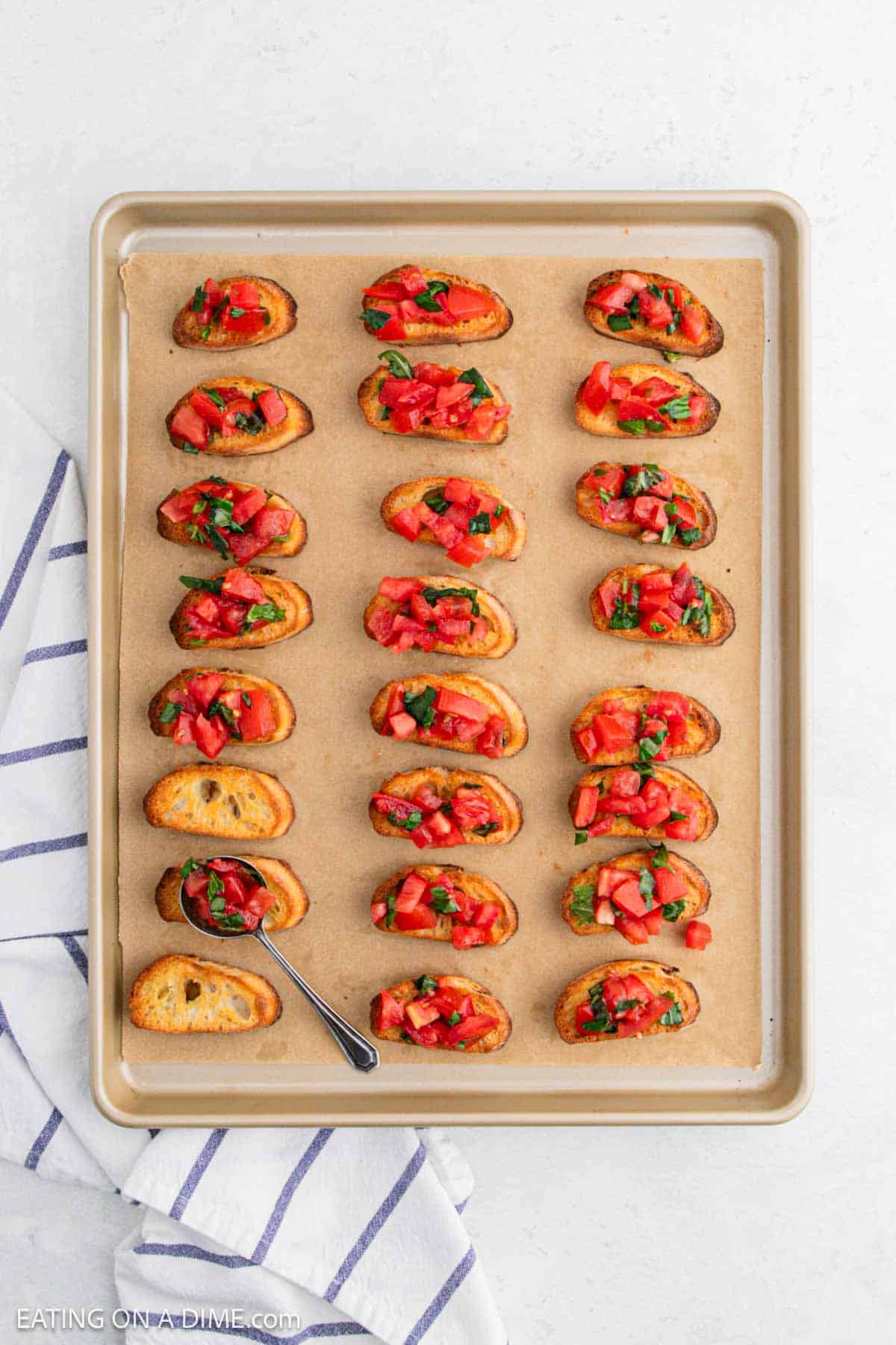 A baking tray with 24 pieces of bruschetta topped with diced tomatoes and herbs. A small spoon rests on the tray's corner. The tray is lined with parchment paper and a striped cloth is visible on the side.