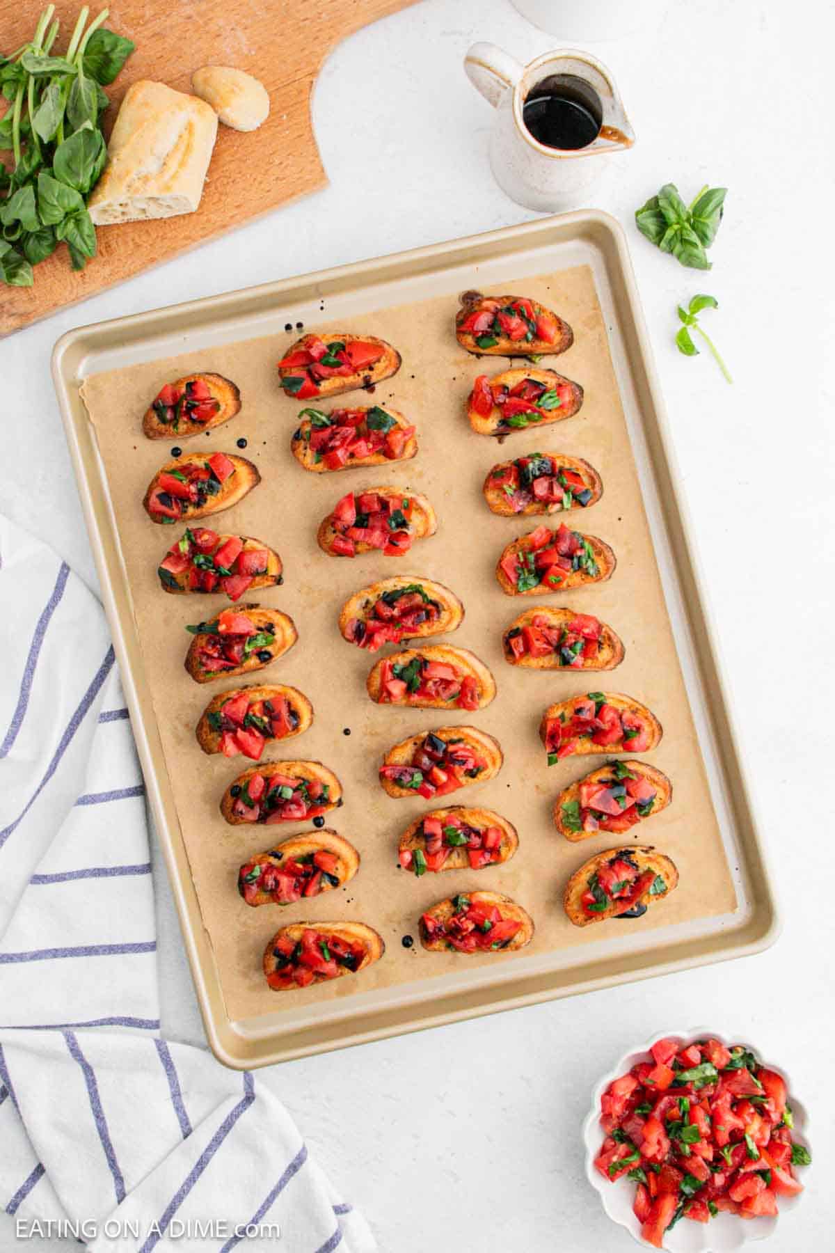 A baking sheet with multiple bruschetta pieces topped with diced tomatoes, basil, and balsamic glaze. Surrounding items include a white sauce jug, a bowl of chopped tomatoes, basil leaves, bread, and a striped towel.