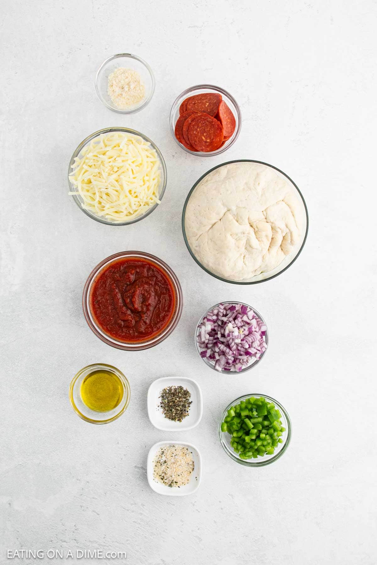 A top view of pizza ingredients on a white surface. Includes a bowl of dough, shredded cheese, tomato sauce, sliced pepperoni, chopped red onion, diced green bell pepper, olive oil, and small bowls of spices and seasonings.