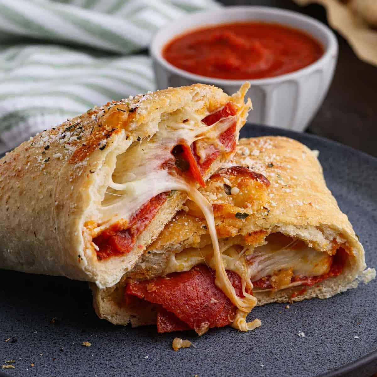 A cheesy pepperoni calzone sliced open, revealing melted cheese and pepperoni inside, is placed on a dark plate. A small bowl of marinara sauce is in the background. A striped cloth is partially visible next to the sauce.