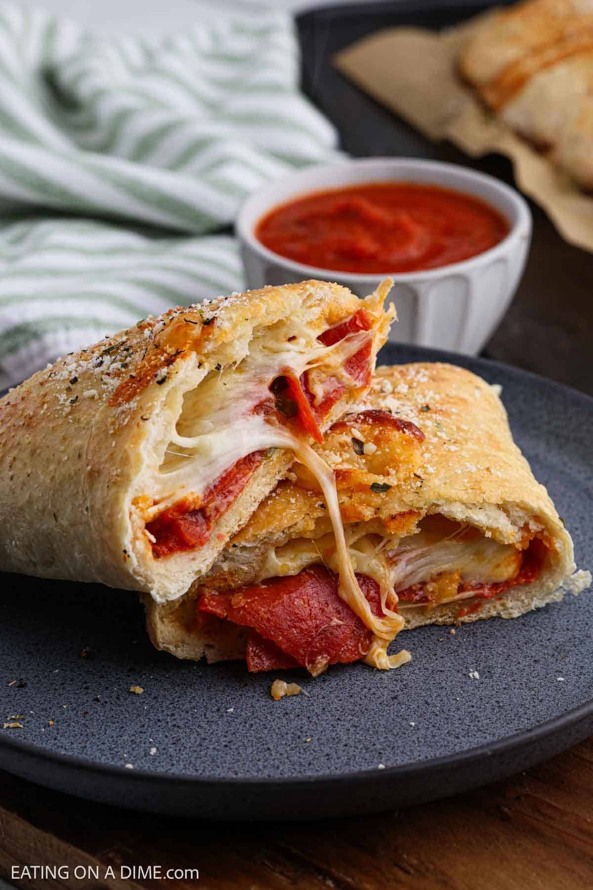 Two slices of stuffed bread with melted cheese and pepperoni are on a dark plate. The bread is golden and sprinkled with seasoning. A small bowl of marinara sauce is in the background, next to a striped green and white cloth.