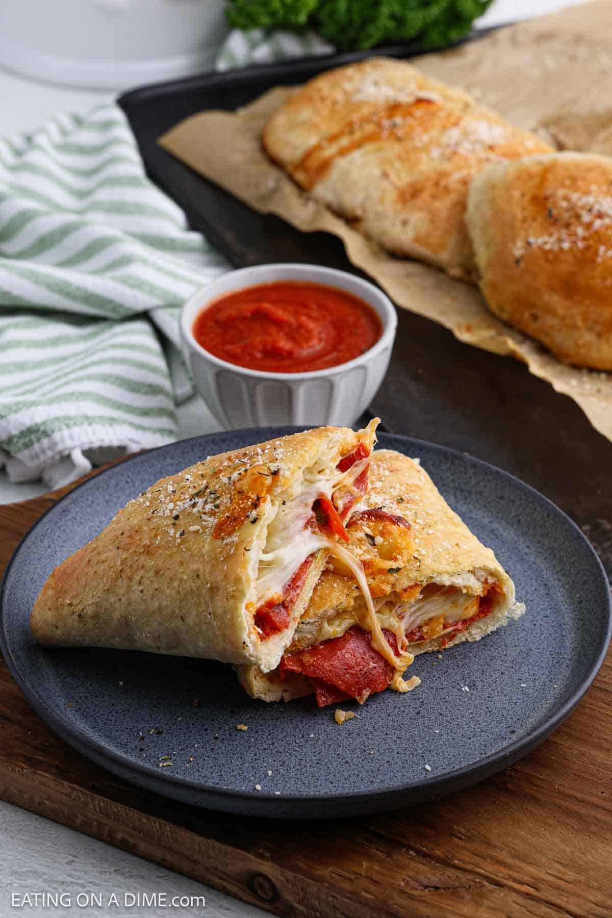 A slice of stuffed calzone on a gray plate, revealing melted cheese and pepperoni. A small bowl of marinara sauce is in the background, alongside the remaining calzone on parchment paper. A green and white cloth is partially visible.