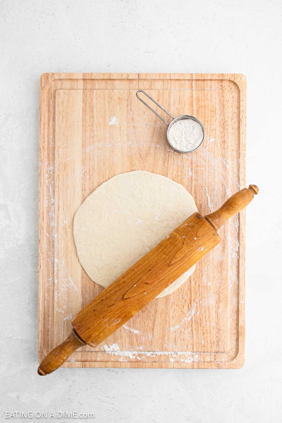 A wooden rolling pin rests on a flat, round piece of dough on a wooden board. A small metal measuring cup with flour is placed near the top edge of the board. The background is a light gray surface.