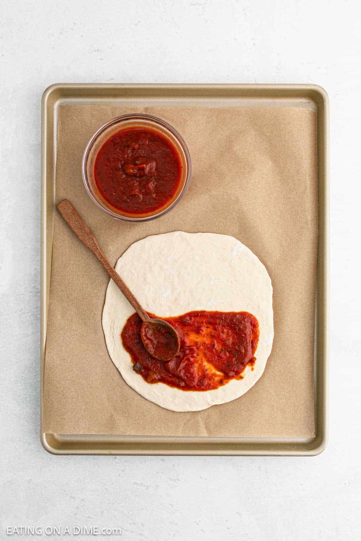 A baking sheet holds a circle of pizza dough on parchment paper, partially covered with tomato sauce. A small bowl of sauce with a wooden spoon rests beside the dough.