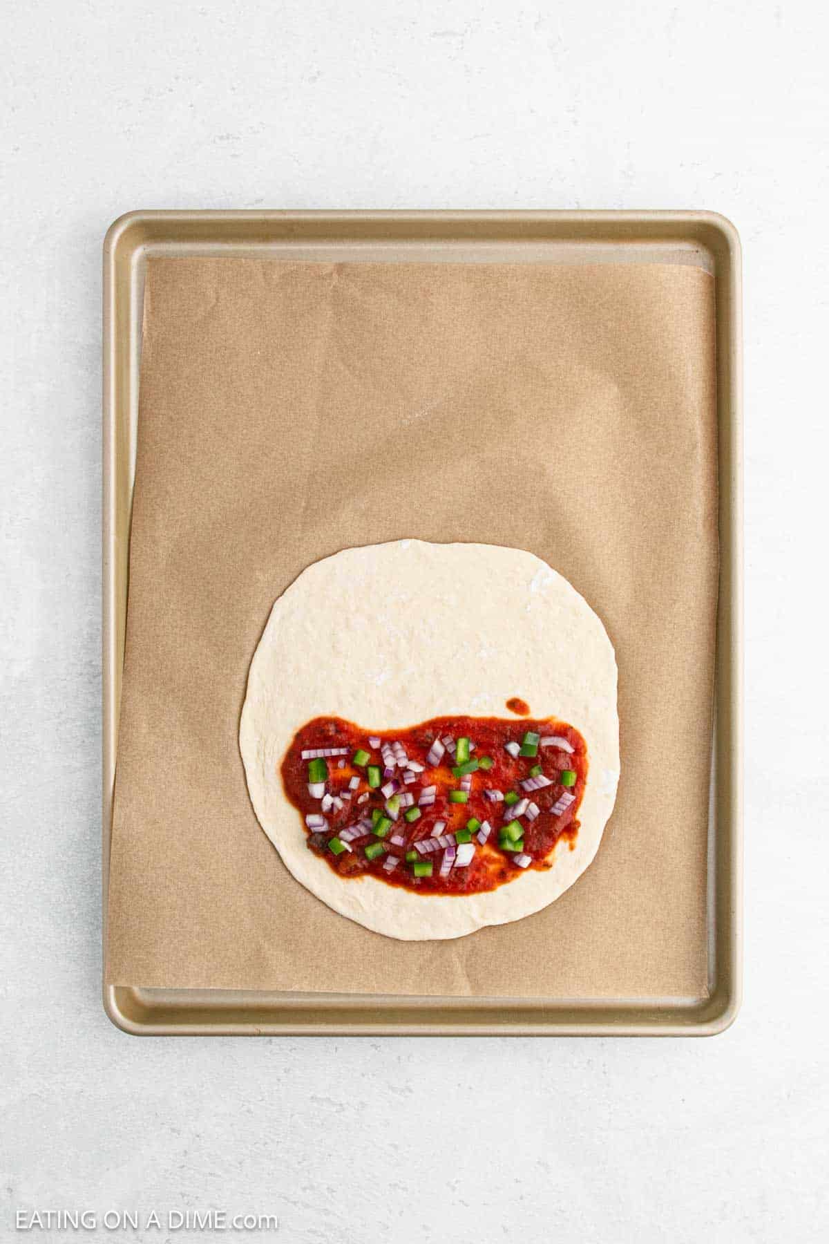 A baking sheet with parchment paper holds a plain tortilla topped with tomato sauce, chopped green bell peppers, and diced red onions. The tortilla is centered on the sheet, ready for additional toppings or baking.