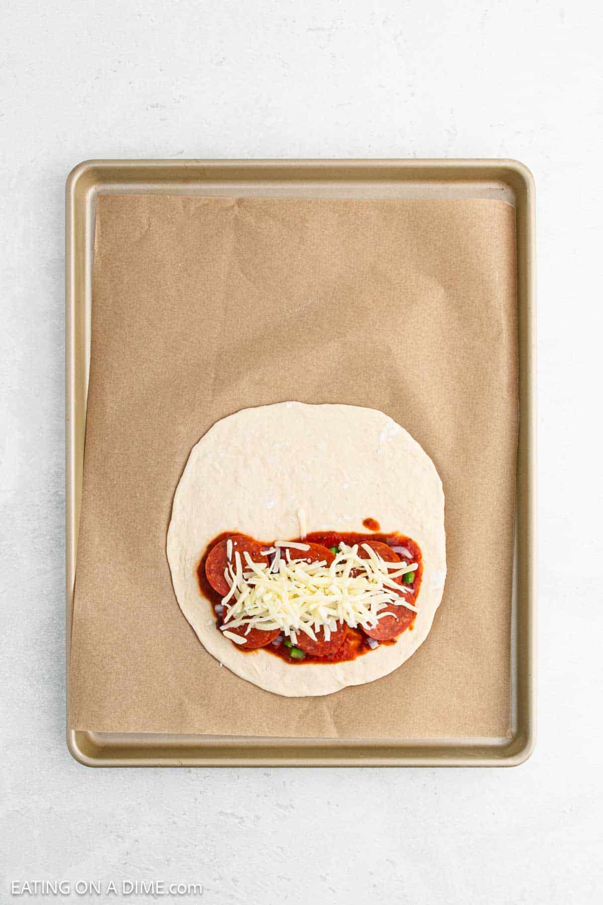 A baking tray lined with parchment paper holds a round piece of dough topped with tomato sauce, pepperoni slices, and shredded cheese, ready to be baked.