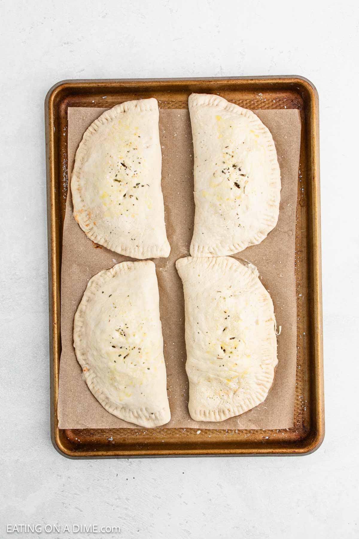 Four uncooked calzones on a parchment-lined baking sheet, sprinkled with seasoning, shown from above.