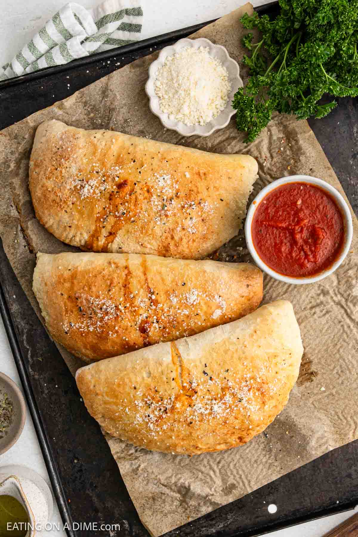 Three golden-brown calzones sprinkled with cheese sit on parchment paper. A small bowl of marinara sauce is beside them for dipping. Fresh parsley is visible in the top-right corner of the baking sheet.