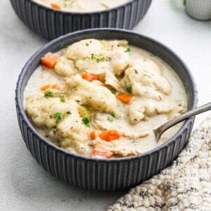 A bowl of creamy chicken and dumplings, part of Meal Plan 46, is garnished with herbs. Visible chunks of chicken, carrots, and dumplings swim in the thick sauce. A spoon rests in the bowl, with a textured cloth subtly framing the cozy scene.