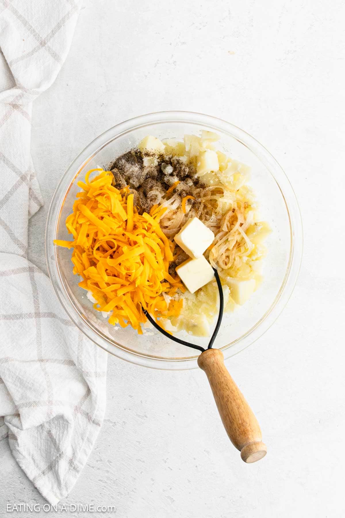 Shredded cheese, slice butter, potatoes, seasoning and cooked onions in a bowl with a potato masher