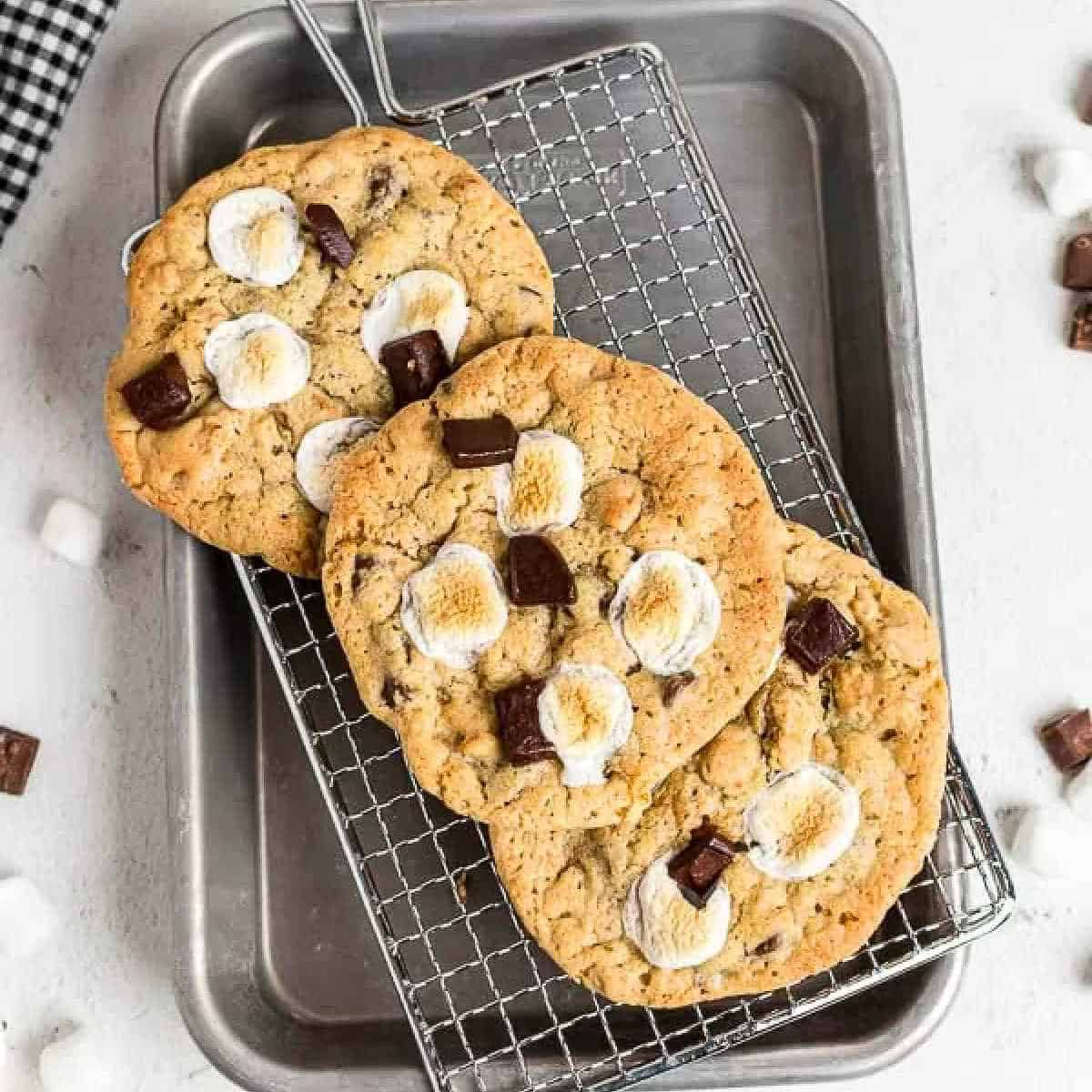 S'mores cookies with toasted marshmallows and chocolate chunks rest on a cooling rack over a baking tray. Scattered mini marshmallows add a playful touch around the tray.