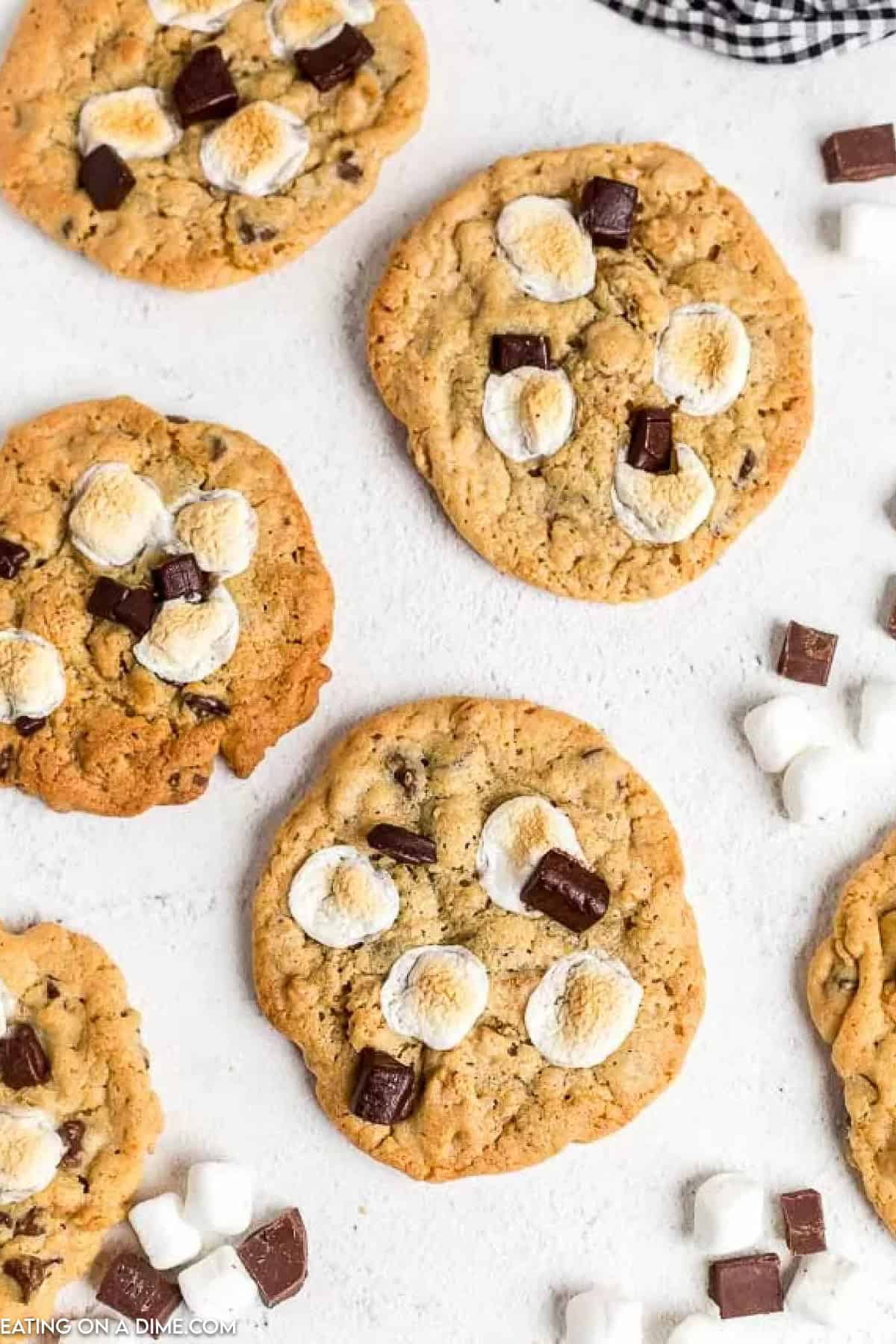 Overhead view of freshly baked s'mores cookies topped with toasted marshmallows and chocolate chunks on a light surface. Scattered around are mini marshmallows and chocolate pieces, with a checkered cloth in the corner.