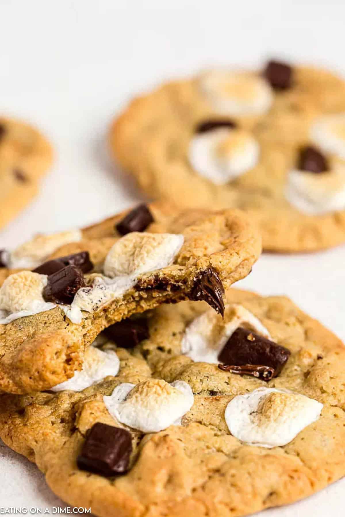 A close-up of S'mores Cookies topped with toasted marshmallows and chocolate chunks. One cookie is broken in half, revealing a soft, gooey interior. These treats rest enticingly on a white surface.